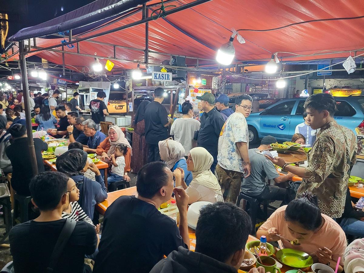 Puesto de comida callejera de noche bajo un toldo rojo, con las mesas corridas llenas de gente cenando